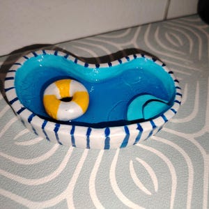 May include: A small, heart-shaped ceramic dish painted to resemble a swimming pool. The pool is filled with blue water and features a yellow and white striped inflatable ring and a blue wave. The rim is white with blue stripes.