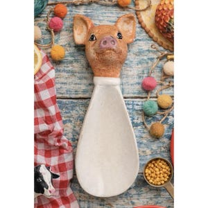 May include: A ceramic spoon with a pig head design. The pig head is light brown with pink details, and the spoon is off-white. The spoon is on a blue wooden surface with colorful pom-poms and a red and white checkered cloth.