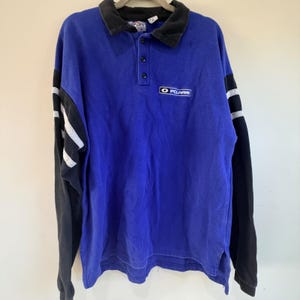 May include: A long-sleeve polo shirt in royal blue with a black collar and black sleeves featuring two horizontal grey stripes. The shirt has a three-button placket and a rectangular patch on the chest with the white text Polaris on a black background.