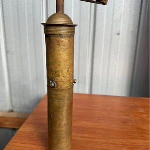 May include: An antique brass pepper mill with a cylindrical body and a hand crank. The mill has a tarnished, aged appearance, with visible wear and tear. The crank handle is made of a lighter-colored metal, and the mill sits on a wooden surface.