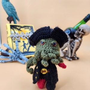 May include: A crocheted green octopus pirate wearing a black pirate hat and red and black clothing. The octopus has red eyes and is holding a sword. The octopus is standing on a beige surface.