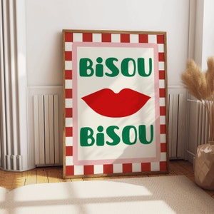 May include: A framed art print with a cream background, featuring a red lip graphic and the word "BISOU" in green, repeated above and below the lips. The print has a pink border and a red and white checkered frame.