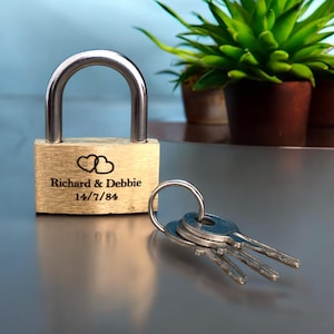 May include: A gold padlock with two keys. The padlock is engraved with the inscription "Richard & Debbie 14/7/84" and two hearts.