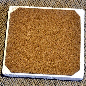 May include: A square cork coaster with a white border. The coaster is made of natural cork and has a slightly rough texture.