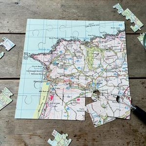 Può includere: Un puzzle raffigurante una dettagliata mappa topografica di una regione costiera con linee di griglia blu, curve di livello e nomi di luoghi. Diversi pezzi sparsi sono posizionati attorno alla mappa parzialmente assemblata e una lente d'ingrandimento.