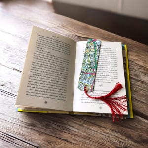 May include: A rectangular bookmark featuring a colorful printed map design with a vibrant red tassel attached to the bottom, resting inside an open book with printed text on the pages. The map displays various street lines and urban grid patterns.