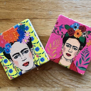 May include: Two square coasters with a colorful floral design and a portrait of Frida Kahlo. The coasters are made of a light pink and yellow material.