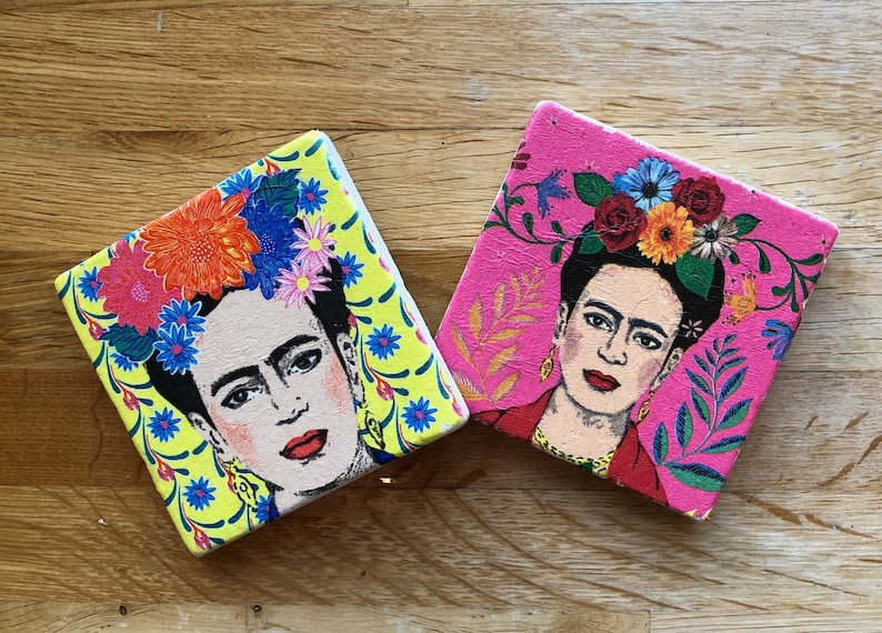 May include: Two square coasters with a colorful floral design and a portrait of Frida Kahlo. The coasters are made of a light pink and yellow material.