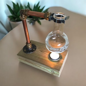 May include: A distinctive lamp with a copper pipe structure and a wooden base. A round glass flask is held by the frame, with a small light source illuminating the flask. The lamp has a rustic, industrial design, with a vintage feel.