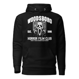 May include: Black hooded sweatshirt with a white graphic. The design includes the text "WOODSBORO HORROR FILM CLUB" and a Scream mask. The text "EST. 1996" is also present. The sweatshirt has a front pocket and a drawstring hood.