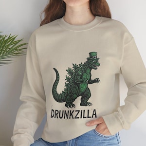 Può includere: Felpa beige con un mostro verde simile a Godzilla con un cappello a cilindro. La grafica include la parola "DRUNKZILLA" in testo nero. La felpa ha maniche lunghe e girocollo.