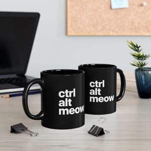 May include: Two black ceramic mugs featuring the white text "ctrl alt meow" printed on the side. Each mug has a curved handle and a cylindrical body. The design is displayed clearly on the front of both mugs, which are positioned on a flat surface.