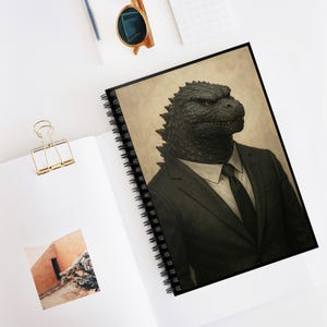 May include: Spiral-bound notebook featuring a detailed illustration of Godzilla in a black suit and tie. The notebook has a black spiral binding and a cream-colored background. The image is a unique and artistic representation of the iconic monster.