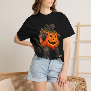 May include: A black short-sleeved t-shirt featuring a graphic print of a grey, scaly monster resembling a dinosaur emerging from a large, orange, carved jack-o'-lantern pumpkin with a green stem and a classic triangular-eyed face design.