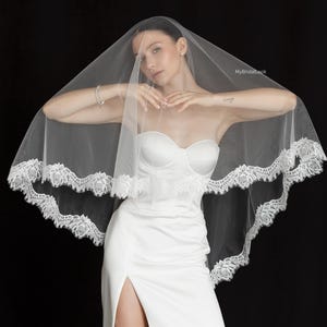 May include: A white wedding veil with a delicate lace trim, draped over a white strapless dress. The veil is sheer, and the lace trim adds a touch of elegance. The dress has a high slit. The background is black.