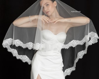 May include: A white wedding veil with a delicate lace trim, draped over a white strapless dress. The veil is sheer, and the lace trim adds a touch of elegance. The dress has a high slit. The background is black.