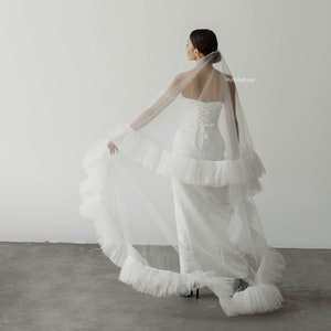 Puede incluir: Una novia con un vestido de novia blanco y un velo largo y fluido. El velo tiene un borde con volantes y está drapeado sobre los hombros de la novia. La novia está de pie en una habitación blanca con una pared blanca detrás de ella.