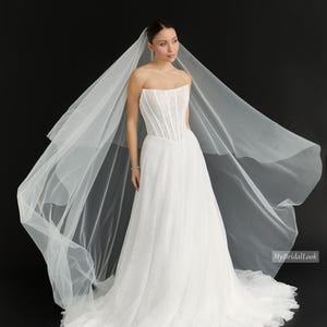 Peut inclure: Une robe de mariée blanche sans bretelles avec un corsage corset structuré et une jupe longue, accompagnée d'un long voile transparent et fluide. Le vêtement présente un motif texturé subtil. Le texte MyBridalLook est visible.