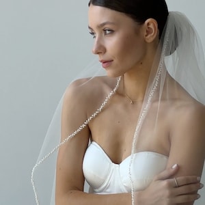 Könnte beinhalten: Ein weißer Braut Schleier mit Perlenbesatz. Der Schleier ist über die Schultern einer Frau gelegt und mit einem Kamm befestigt.