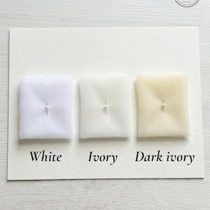 May include: A display of three fabric swatches in white, ivory, and dark ivory, each pinned with a small pearl. The swatches are arranged on a white card, with the color names printed below each sample. A logo is in the upper right corner.
