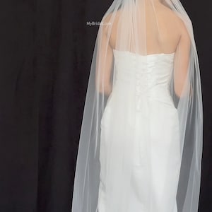 May include: A white wedding dress with a long, flowing veil. The dress has a fitted bodice and a simple design. The sheer veil drapes over the shoulders and back. The background is black.