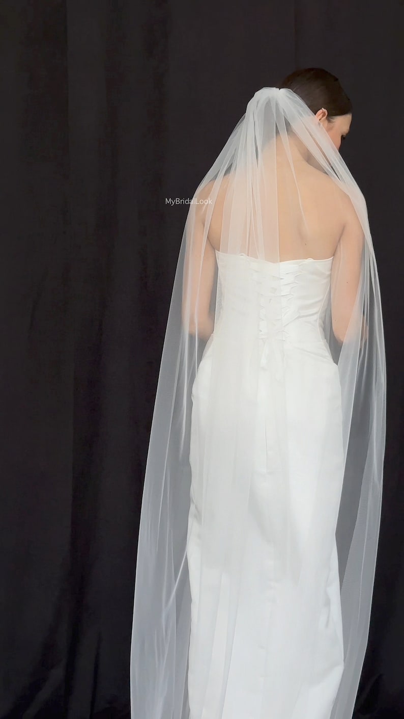 May include: A white wedding dress with a long, flowing veil. The dress has a fitted bodice and a simple design. The sheer veil drapes over the shoulders and back. The background is black.