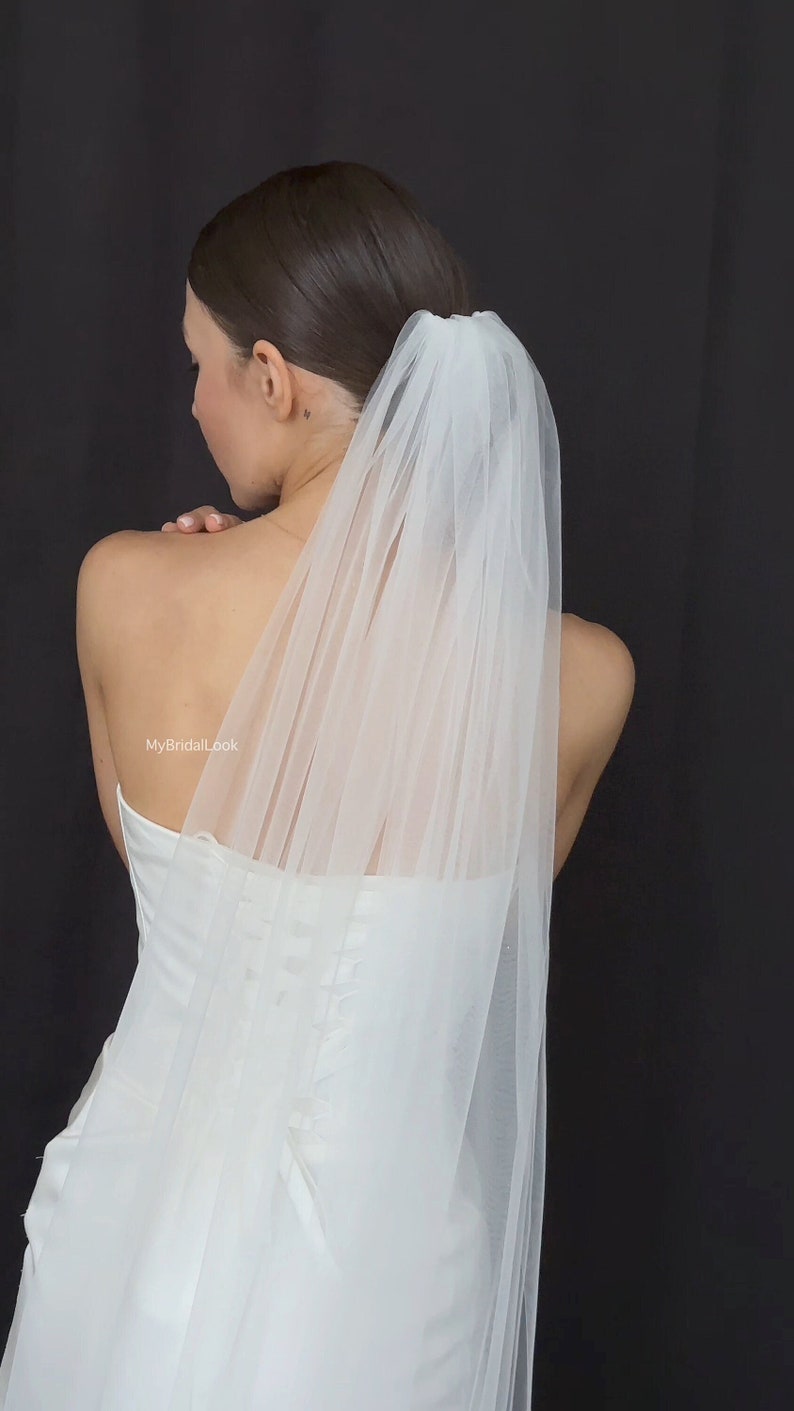 May include: A white bridal veil, made of sheer fabric, falls down the back. The veil is attached to the hair and worn with a white strapless wedding dress. The background is a solid black.