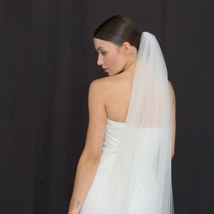 May include: A bride in a white strapless wedding dress and a sheer veil. The veil is attached at the back of the head and falls down the back. The background is a solid black.