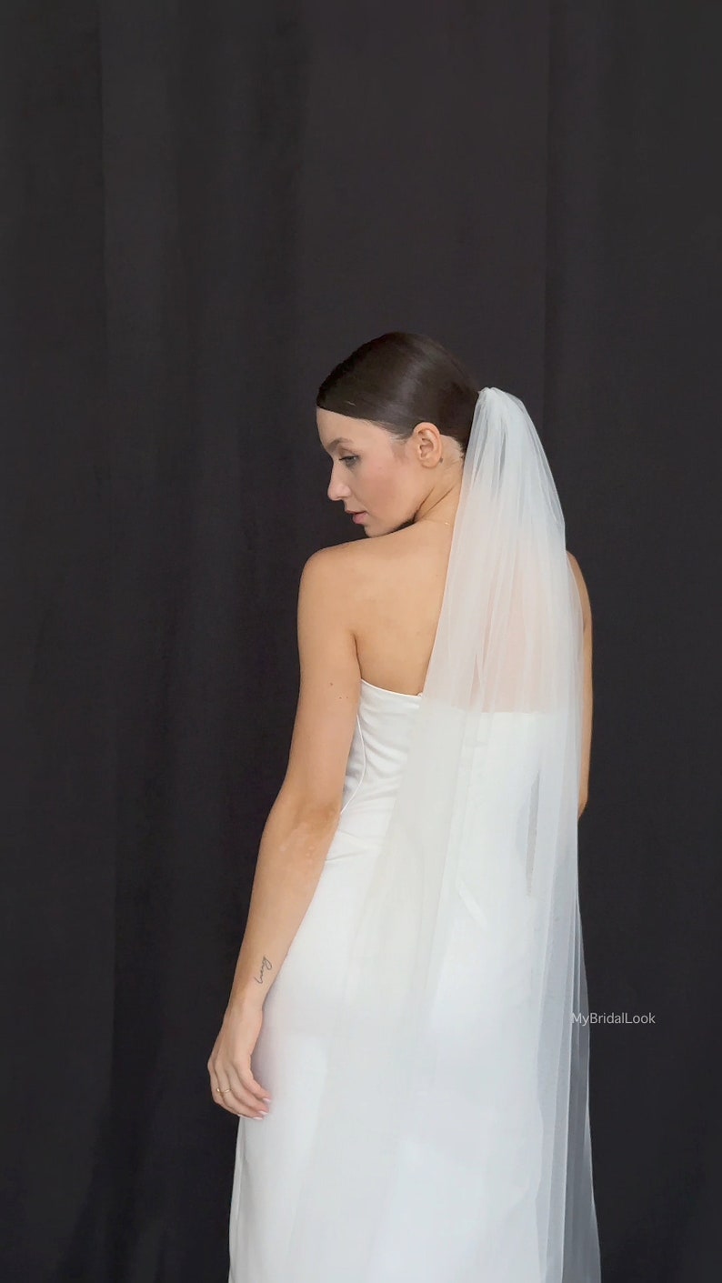 May include: A bride in a white strapless wedding dress and a sheer veil. The veil is attached at the back of the head and falls down the back. The background is a solid black.