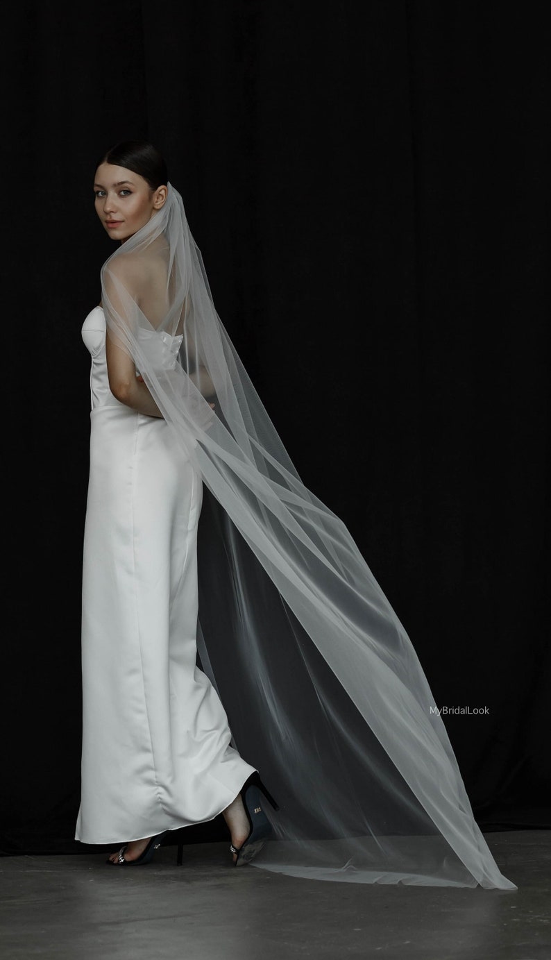 May include: A white bridal jumpsuit with a long, flowing veil. The veil drapes down the back, creating a dramatic effect. The outfit is simple and elegant, ideal for a wedding. The background is black.