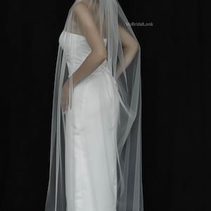 May include: A bride in a white strapless wedding dress with a long, sheer veil. The veil is attached at the back of the head and drapes down the back. The background is black, and the image is from MyBridalLook.