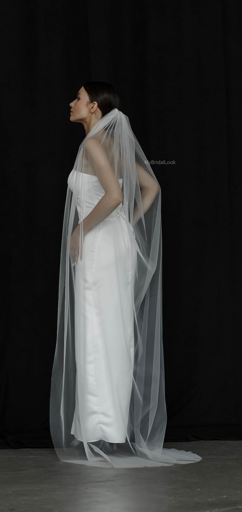 May include: A bride in a white strapless wedding dress with a long, sheer veil. The veil is attached at the back of the head and drapes down the back. The background is black, and the image is from MyBridalLook.