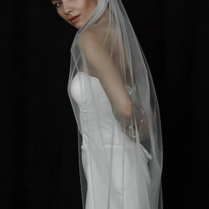 May include: A bride in a white strapless wedding dress and sheer veil against a black backdrop. The veil drapes over the shoulders and back. The dress is fitted and simple. The image is a studio shot.