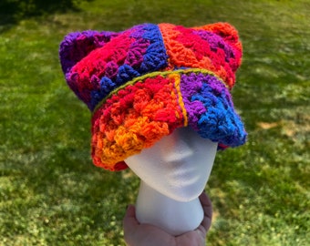 May include: A colorful crocheted hat with a rainbow pattern. The hat is made with different shades of red, orange, yellow, green, blue, and purple yarn. The hat has a unique design with a pointed top and a wide brim.