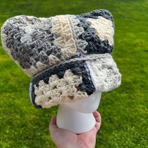 May include: A crocheted hat with a patchwork design in shades of gray, white, and cream. The hat is made with a granny square stitch and has a floppy brim.