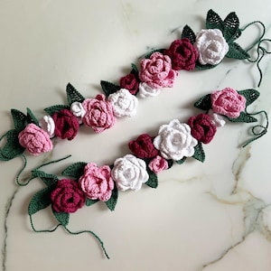 Op de afbeelding: Een handgemaakte krans van gehaakte bloemen in tinten roze, wit en bordeauxrood. De krans is gemaakt met groene bladeren en heeft een rustieke, vintage uitstraling.