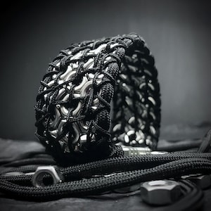 Könnte beinhalten: Ein schwarzes und silbernes Armband aus miteinander verwobenen Metallringen und schwarzem Band. Das Armband ist ein einzigartiges und stilvolles Accessoire.