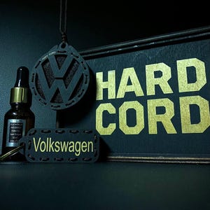 May include: Black car air freshener with a Volkswagen logo, a small tag with "Volkswagen" in gold lettering, and a wooden sign that says "HARD CORD" in gold lettering. A small brown bottle with a dropper is also in the image.