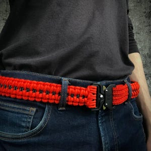Può includere: Una cintura in paracord rosso e nero con una fibbia in metallo nero. La cintura è indossata in vita da una persona che indossa jeans blu.
