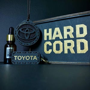 May include: Black accessories with gold lettering, including a Toyota keychain, a Hard Cord sign, and a car air freshener with the Toyota logo. A small bottle with a dropper is also present.