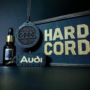 Könnte beinhalten: Ein dunkles Auto-Lufterfrischer-Set mit einem Audi-Logo-Anhänger, einem Schlüsselanhänger und einer Flasche ätherischen Öls. Auf dem Schild steht "HARD CORD" in goldfarbenen Buchstaben. Der Hintergrund ist dunkel.