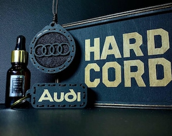 May include: A dark-colored car air freshener set featuring an Audi logo pendant, a keychain, and a bottle of essential oil. The sign reads "HARD CORD" in gold lettering. The background is dark.