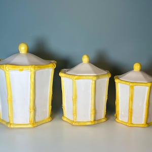 May include: A set of three white ceramic canisters with yellow bamboo-style trim. Each hexagonal container features a matching removable lid with a small yellow knob handle. The canisters are arranged in descending order of size from left to right.