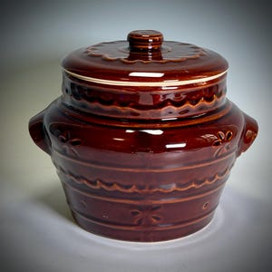May include: A brown ceramic lidded jar with a glossy finish. The jar has a round shape with two handles and decorative floral and wavy patterns. The lid has a scalloped edge and a small knob. A classic kitchenware item.