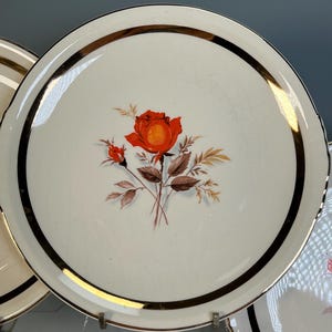 May include: A white ceramic plate with a gold rim and a black accent band. The plate features a red rose and two buds with brown and gold leaves. The plate is part of a set of vintage dinnerware.