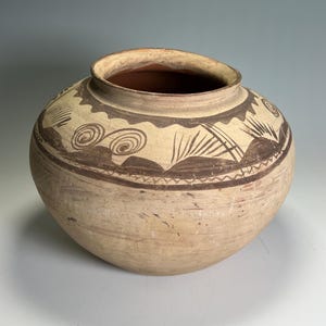 May include: A beige ceramic pot with a wide opening and rounded body. The pot features brown geometric patterns around the rim and a band of decorative elements. The interior is a darker brown color.