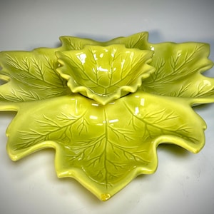 May include: A three-piece serving set in a leaf design. The set includes two large leaf-shaped platters and a smaller bowl, all in a glossy, light green color. The leaf veins are detailed, adding texture to the ceramic pieces.