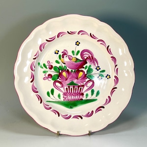 May include: A round ceramic plate with a scalloped edge, featuring a hand-painted design of a pink rooster perched atop a pink basket filled with green leaves and pink flowers, surrounded by a border of purple brushstroke accents on white.