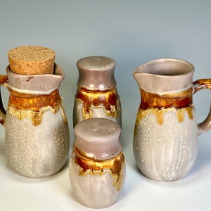 May include: A ceramic pitcher with a cork stopper, two salt shakers, and a creamer. The set features a speckled white glaze with brown and gold accents. The pitcher and creamer have handles.