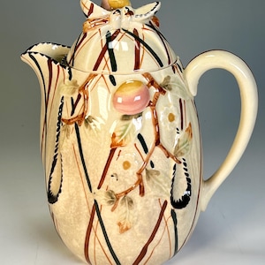 May include: Cream-colored ceramic teapot with a lid, handle, and spout. The teapot is decorated with brown, black, and green vertical and diagonal lines. It features a branch with pink and yellow fruit and green leaves.